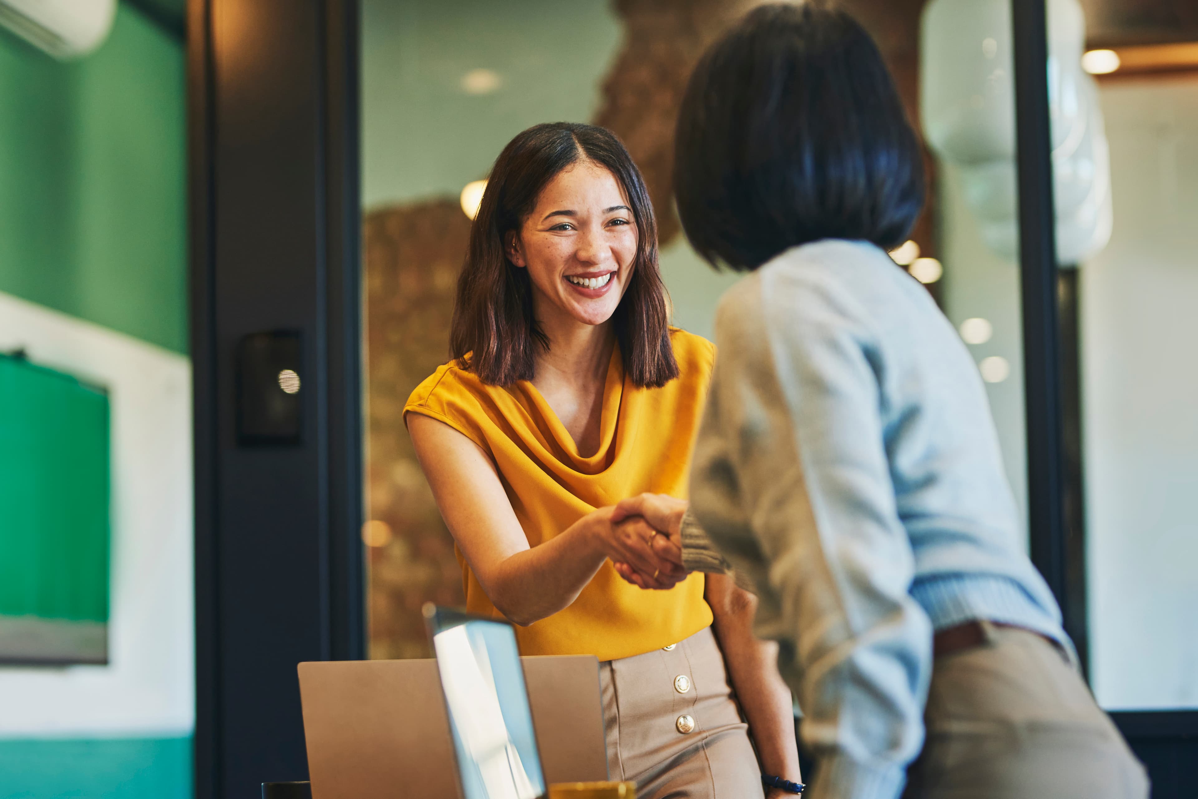 Two women in an office setting are shaking hands and smiling, one wearing a yellow top and the other in a gray sweater. A laptop is on the table.